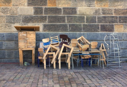 An outdoor scene featuring a collection of wooden furniture pieces and metal chairs arranged against a weathered brick wall, situated on a cobblestone pavement. The wooden furniture includes chairs and small tables, some with light-colored unfinished wood and others stained darker. The chairs are stacked or leaning, with visible grain and texture in the wood. To the left, there is a wooden crate or box with a slatted design, slightly taller than the surrounding furniture, positioned on the ground. On the right side, there are a few metal chairs with a wire frame design, leaning against the brick wall. The overall environment suggests an outdoor storage or disposal area for furniture, relevant to rubbish removal services in Edmonton, with natural daylight illuminating the scene and highlighting the textures and colors of the materials.