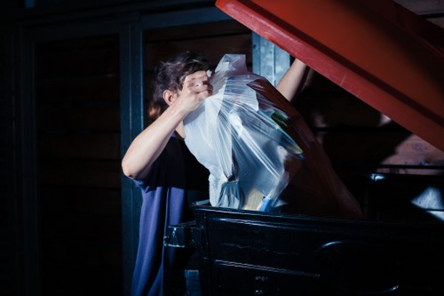 A person, partially visible with short dark hair, is seen placing a clear plastic bag filled with various types of rubbish into a large black skip or wheelie bin outdoors. The bin is positioned on a paved surface, with a red lid partially open and resting at an angle above the bin. The background shows a wooden fence or shed with vertical dark brown planks, and metal supports painted blue. The lighting suggests an overcast or indoor environment, with the focus on the action of rubbish disposal. This scene is typical of commercial waste collection, emphasizing proper waste management practices, and is relevant to local rubbish removal services in Edmonton, underscoring the environmentally responsible disposal of waste materials.