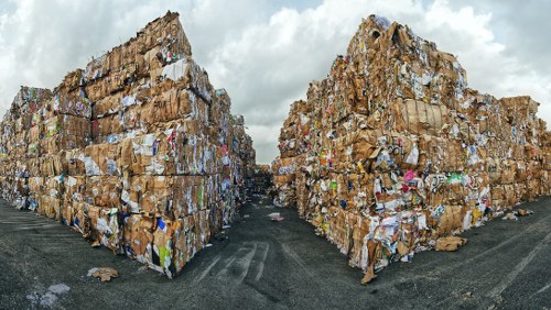 Two large, tightly stacked piles of mixed waste paper and cardboard are situated outdoors on a gravel or paved surface, with a narrow pathway between them. The waste materials include various corrugated cardboard boxes, some with printed labels or logos, and flattened sheets of paper. The cardboard displays a range of colours from brown to faded white, with some pieces showing signs of weathering or slight damage. The background features an overcast sky with grey clouds, creating diffuse natural lighting that highlights the textures and details of the waste. The piles are positioned on either side of the pathway, suggesting they are part of a waste collection or disposal area, possibly associated with commercial waste management services such as those provided by Commercial Waste Edmonton, serving the Edmonton area. The overall scene conveys an industrial waste storage context relevant to rubbish removal and waste management operations in a local urban environment.