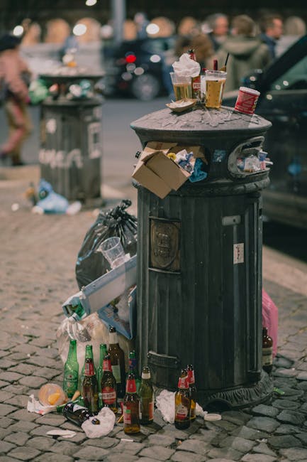 A black wheeled rubbish bin with the label 'ST. JOHN'S' positioned on a paved sidewalk near a curb during nighttime. The bin is open, revealing a mixture of discarded paper, cardboard, and plastic mat